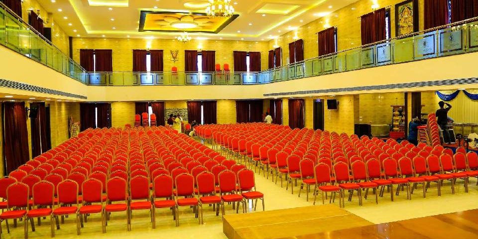 A wedding hall with hundreds of red coloured chairs arranged for guest seating, ceiling lights and decor beautify the hall.