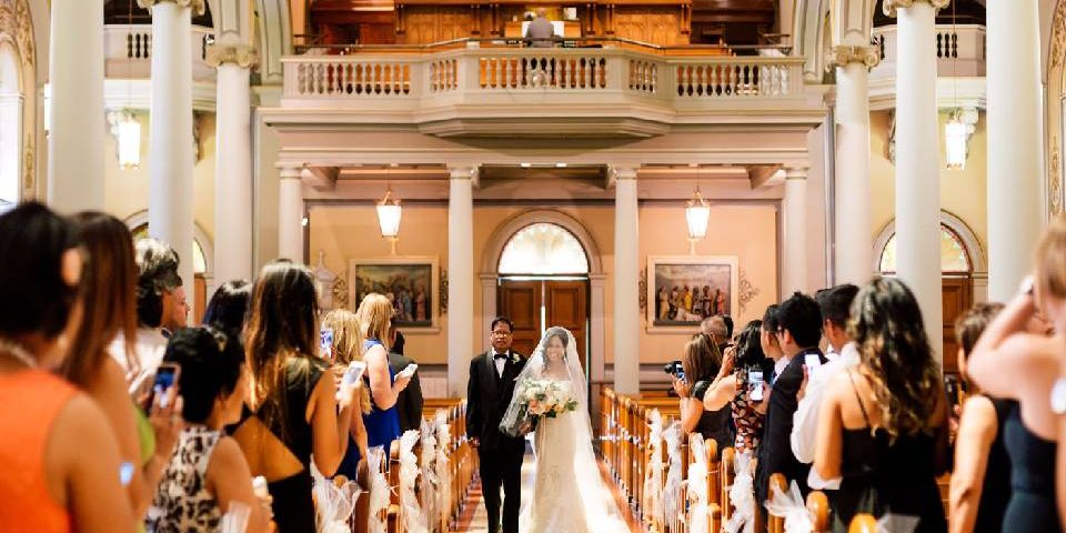 A Christian bride with her father walking into the church for her wedding and guests looking at her.